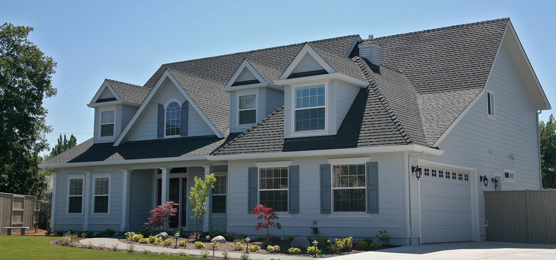 A two-story residential house with white siding, black roofing, and a gray garage door. The house features multiple windows, a central front door, and a porch with a railing.