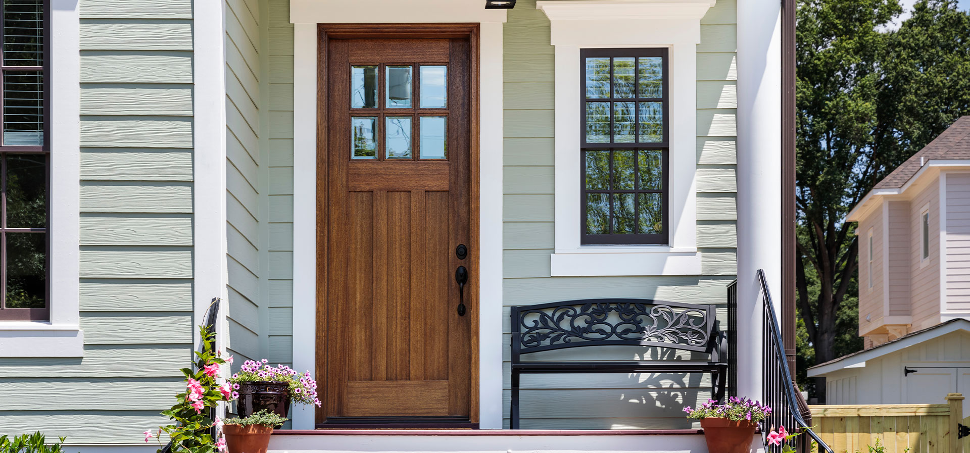 The image shows a two-story house with a white exterior, a black front door with a window above it, and a porch with a bench. There is a wooden front gate, and the house number is visible on the mailbox.
