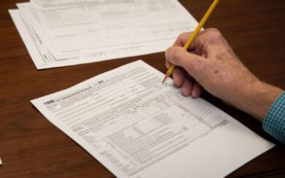 The image depicts a person s hand holding a pencil over a paper with printed text, suggesting the act of filling out a form, likely a tax return given the context of the papers and the setting which appears to be an office environment.